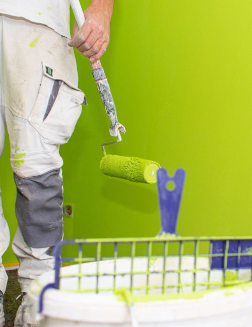 Gebäude- und Hausmeisterservice, Malermeister, Renovierung Ein Maler bei Renovierungsarbeiten mit Malerrolle, Farbeimer und abgeklebter, grüner Wand